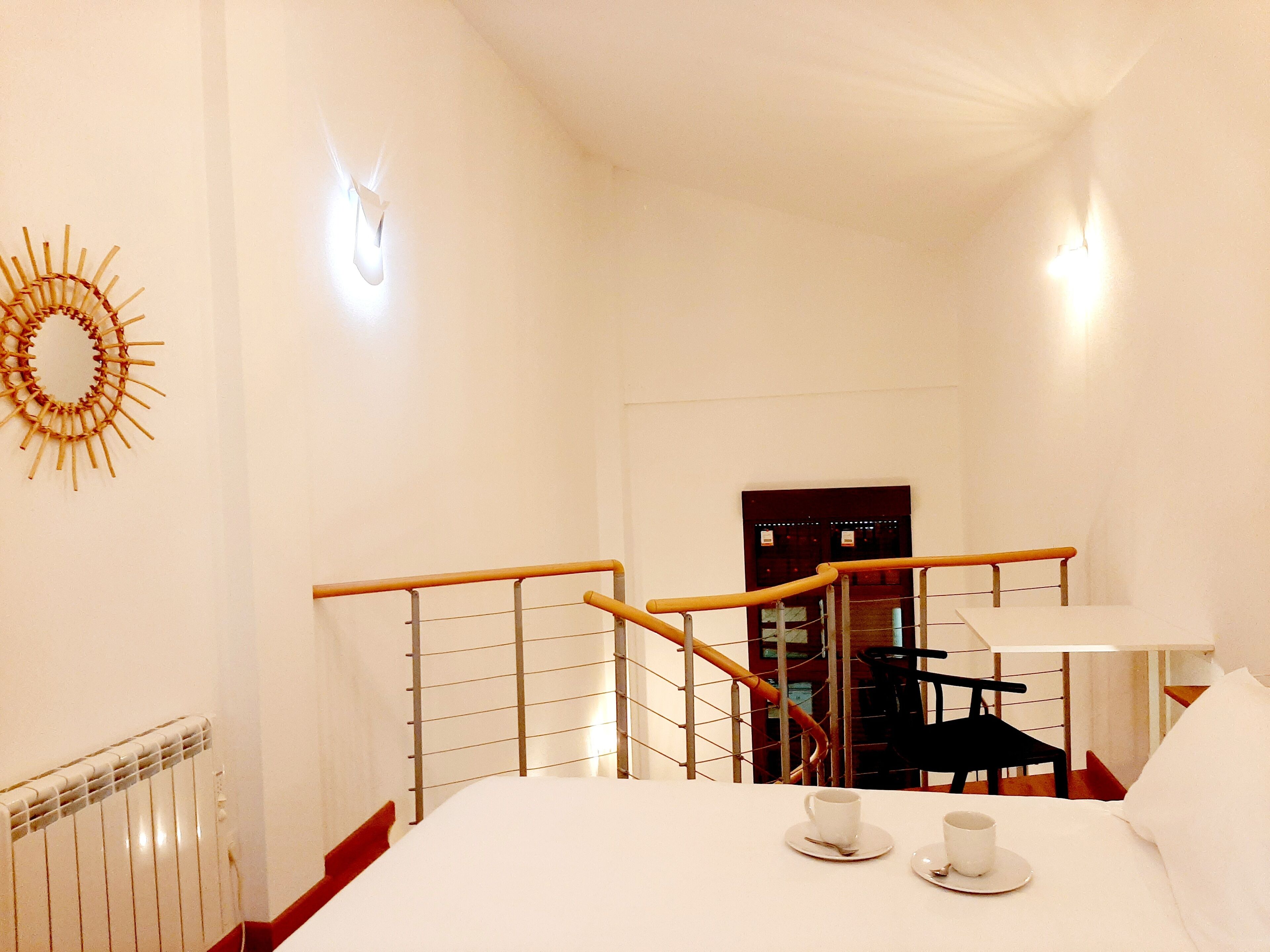Una habitación de hotel con una cama, una mesa de desayuno con dos tazas y platillos, una silla de madera, una silla negra, un radiador blanco, un suelo de madera y un aplique de pared.
