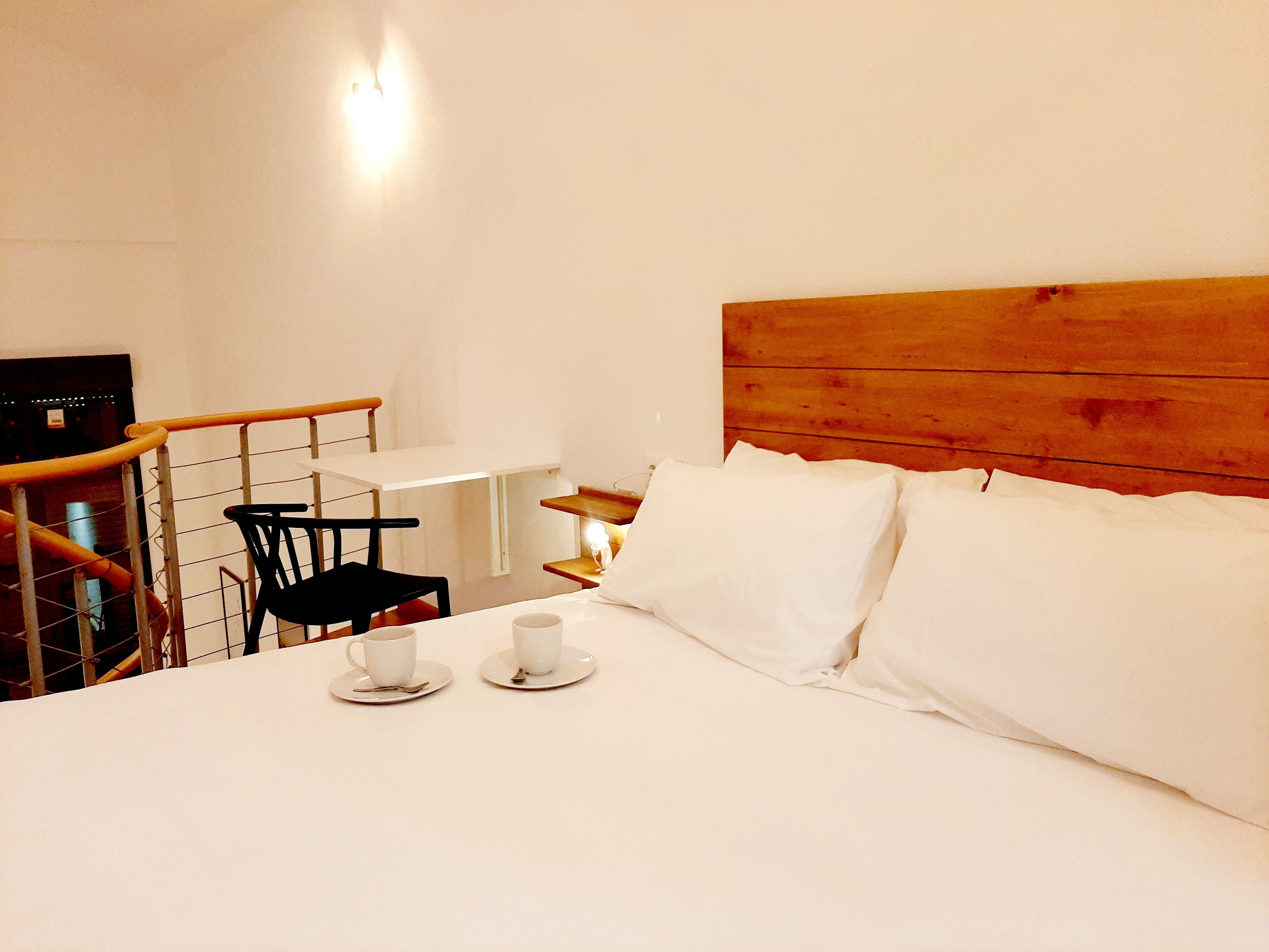 A hotel room with a wooden headboard, a bed with white linens, a small table with a chair, and two cups on the bed.
