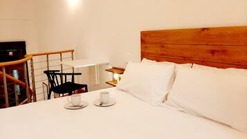 A hotel room with a wooden headboard, a bed with white linens, a small table with a chair, and two cups on the bed.