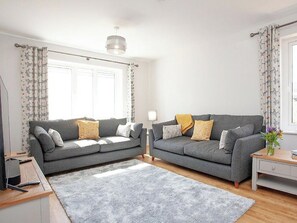 A living room with two grey sofas, a grey rug, a wooden side table, and a window with curtains. - 5 bedroom accommodation in Crantock, near Newquay (Crantock, near Newquay)
