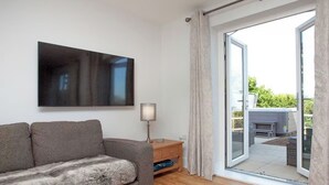 Una sala de estar moderna con un sofá gris, un televisor de pantalla plana, una lámpara de pie y un ventanal corredizo que da a un espacio exterior.