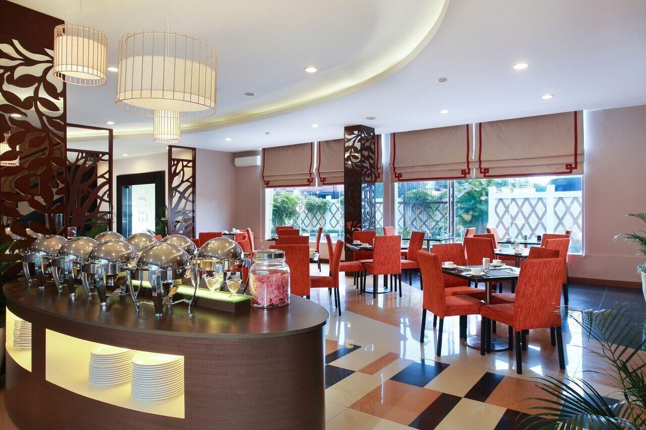 A hotel lobby with a reception desk, multiple dining tables with red chairs, large windows with beige curtains, and a decorative wall with a leaf pattern.