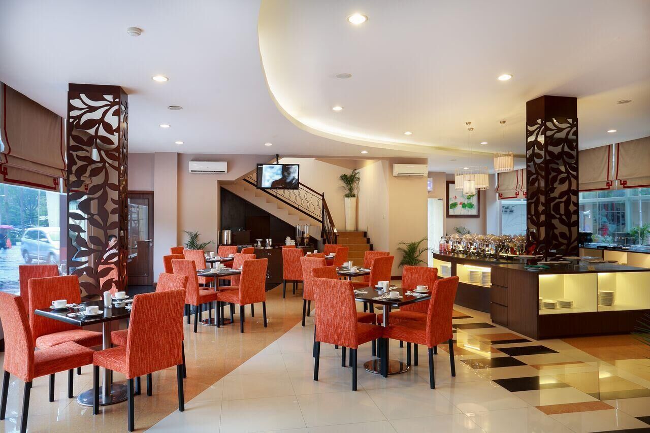A hotel lobby with a central staircase, multiple dining tables with orange chairs, a bar area, and decorative panels.
