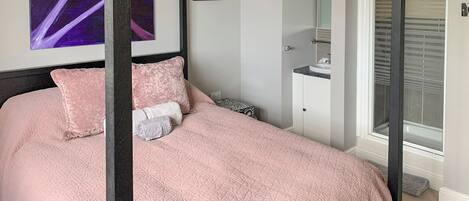 A bedroom with a four-poster bed, a nightstand, a bedside table, a window with blinds, and a door leading to another room.