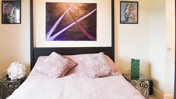 Une chambre à coucher avec un grand lit, deux oreillers, un tableau abstrait encadré et une petite table sur laquelle est posé un objet décoratif.