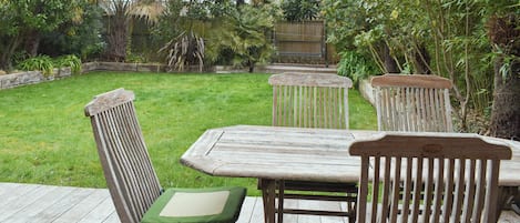 A wooden deck with a table and chairs, surrounded by a grassy yard and various plants.