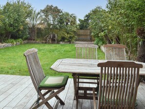A wooden deck with a table and chairs, surrounded by a grassy yard and various plants.