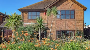 A two-story brick house with a gabled roof, surrounded by a garden with orange lilies and palm trees.