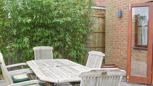 Une table en bois pour l’extérieur, avec quatre chaises, un mur en briques et une plante en bambou en arrière-plan.