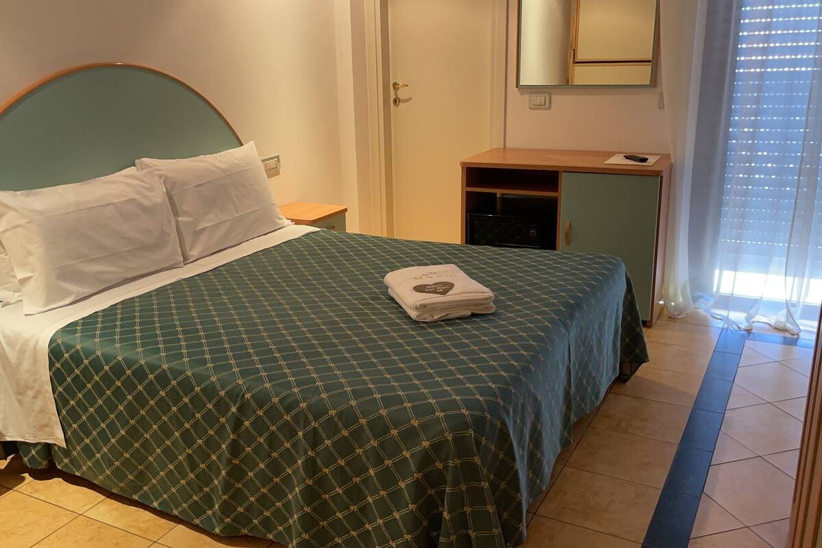 Cama bem arrumada com colcha verde estampada e almofadas brancas, mesa de cabeceira de madeira, um pequeno armário de madeira e um espelho na parede.