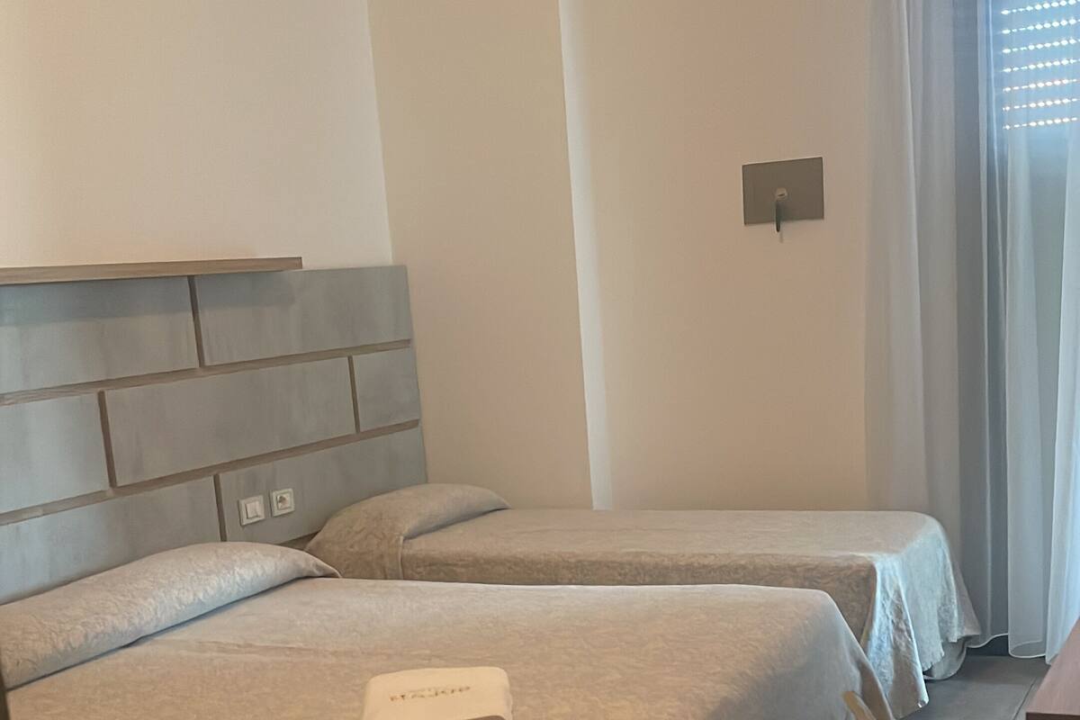 Quarto de hotel com duas camas, uma cadeira e uma janela com cortinas.