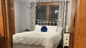 Quarto com piso de madeira, cama branca com travesseiro azul, mesa de cabeceira com abajur e janela com cortinas estampadas.