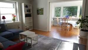 Ein Wohnzimmer mit einem blauen Sofa, einem weißen Couchtisch und einem Bücherregal. Es gibt einen Essbereich mit Tisch und Stühlen sowie einen Blick auf Grünflächen außerhalb.