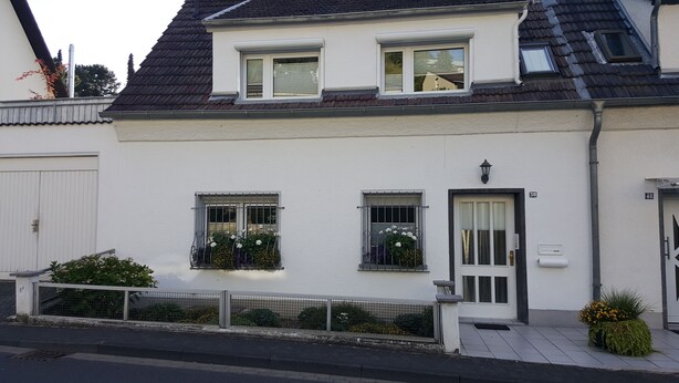 Ein zweistöckiges Wohngebäude mit weißer Fassade, zwei Fenstern mit Gitterstäben, einer Tür mit Klingelschild und einer kleinen Grünfläche mit Pflanzen.