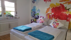 Ein Schlafzimmer mit zwei Betten, einem Fenster mit Blick auf Grünflächen, einem Heizkörper und einem farbenfrohen Wandgemälde mit Blumenmuster.