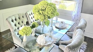 A dining area with a glass-top table, upholstered chairs, a chandelier, and a view through a window with patterned curtains. - ***OASIS*** (Ingersoll)