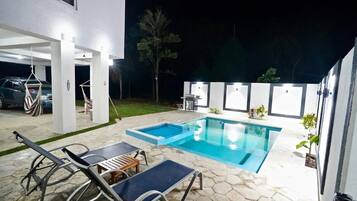 A swimming pool with lounge chairs and a small table, surrounded by stone tiles and a white building.