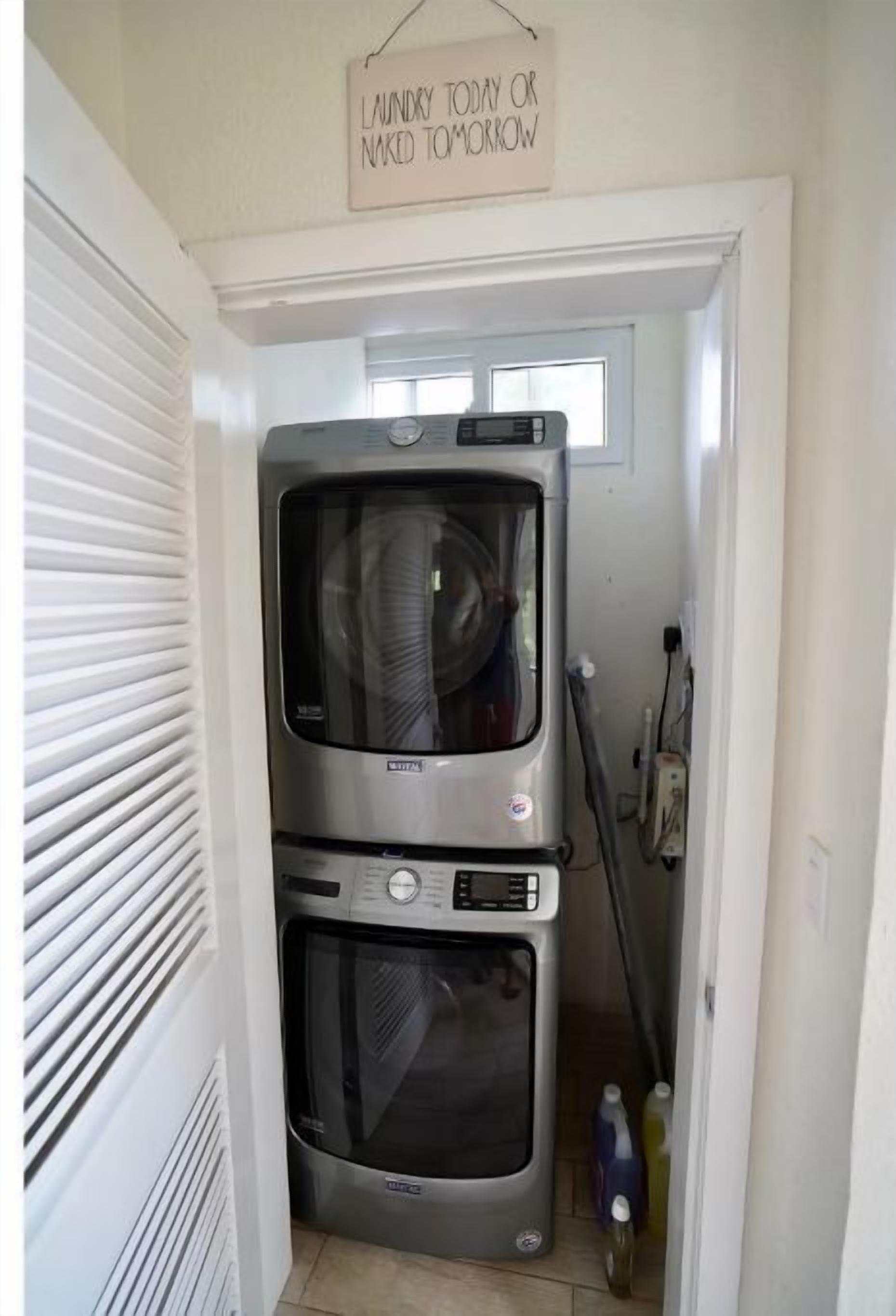 Een kleine wasruimte met een wasmachine en droger boven elkaar, een bord met de tekst 'Was vandaag of wandel morgen naakt' en een dweil die tegen de muur staat.