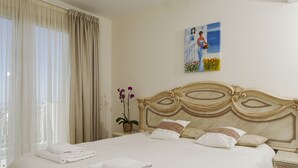 A neatly made bed with white linens and pillows, a potted plant, and a painting on the wall.