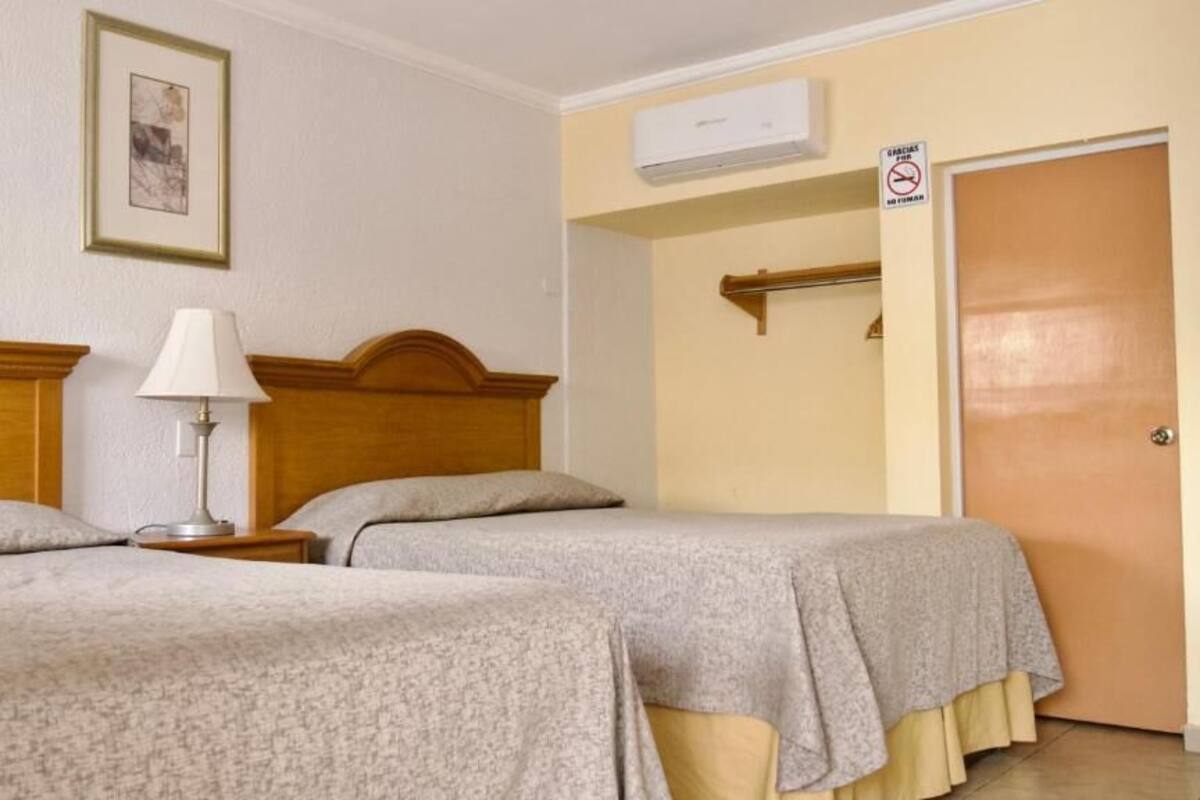 A hotel room with two beds, a bedside lamp, a framed picture on the wall, an air conditioning unit, and a door with a no smoking sign.
