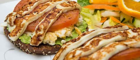 Un sándwich de pollo a la parrilla, aguacate y tomate sobre un pan con semillas, acompañado de una ensalada y una rodaja de naranja.