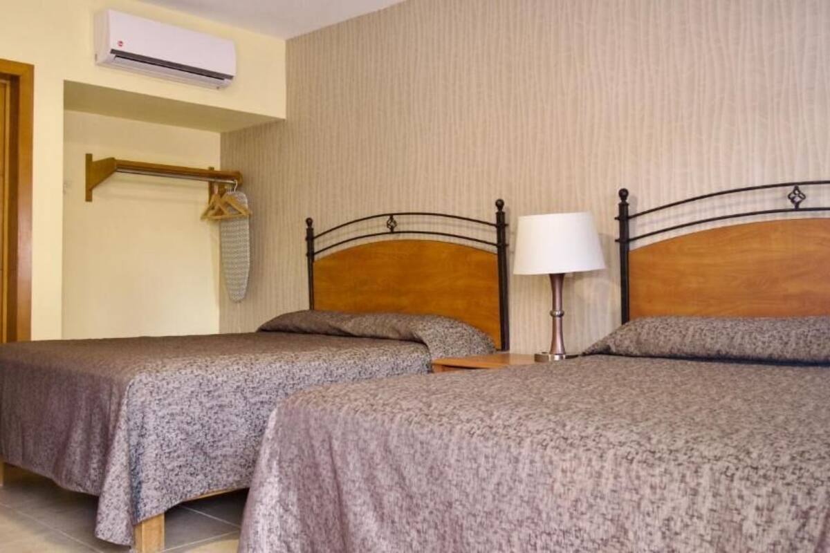 A hotel room with two beds, a wooden headboard, a bedside table with a lamp, a hanging coat rack, and an air conditioning unit.