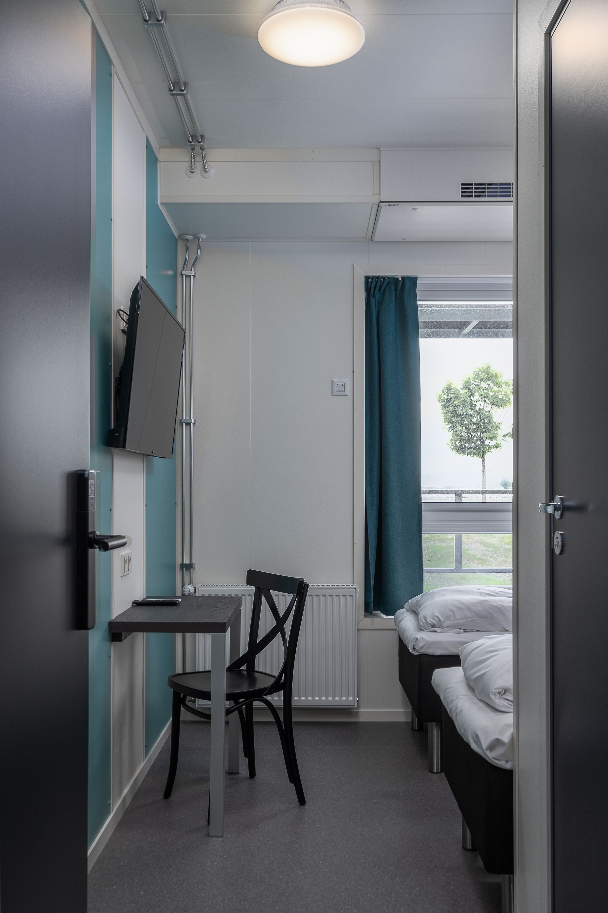 A compact room with a bed, a desk, and a chair. There is a window with a curtain, a radiator, and a mounted television.