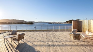 A wooden deck with wicker furniture and a view of a body of water and distant land. - Aiden By Best Western Trondheim Airport (Stjordal)