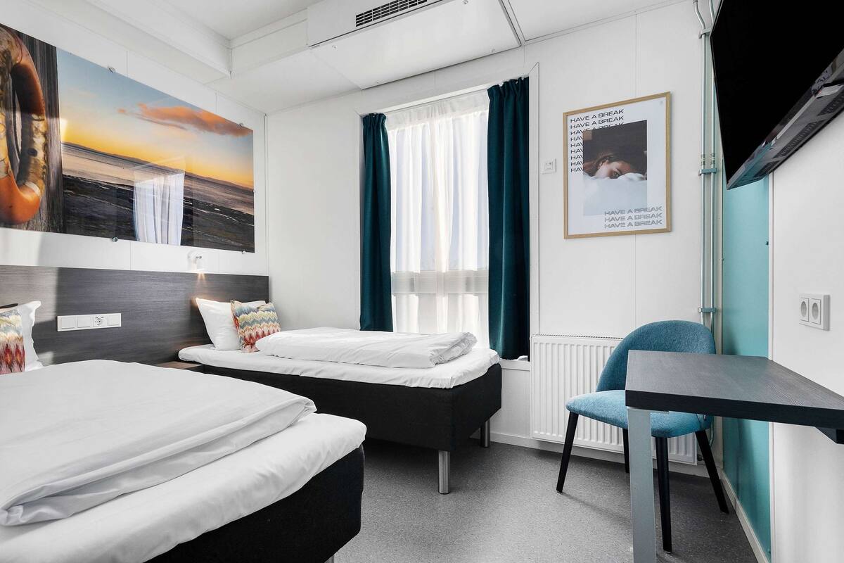A modern hotel room with two beds, a desk, a chair, and a large painting of a sunset.