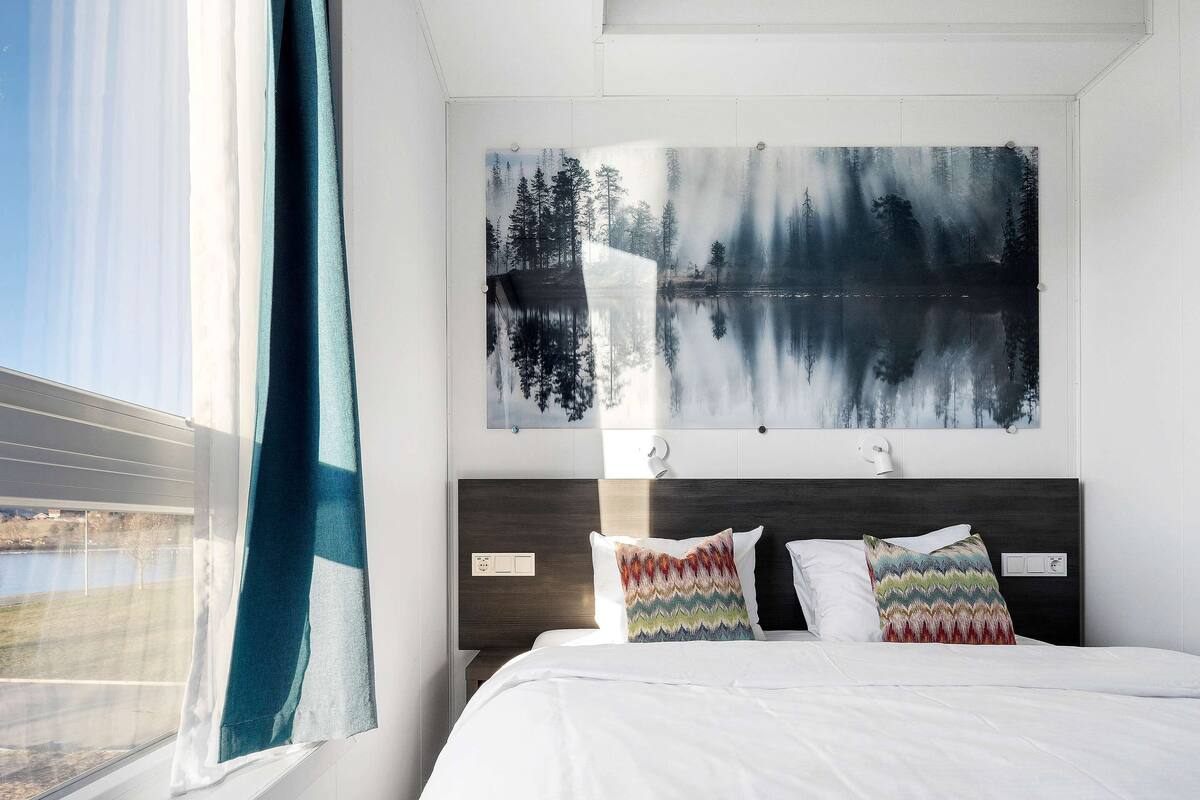 A bedroom with a bed, a window with curtains, and a painting of a forest and lake on the wall.