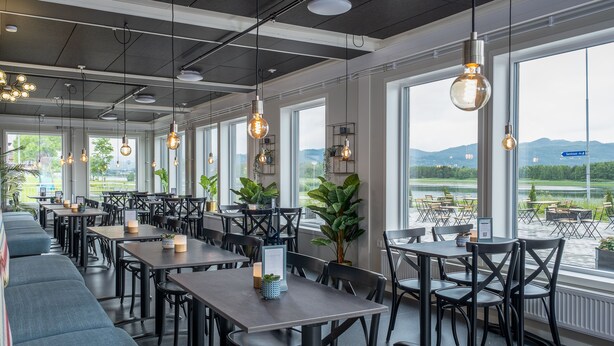 A modern restaurant with high ceilings, exposed ductwork, and large windows offering views of a lake and mountains. Tables and chairs are arranged for dining. - Aiden By Best Western Trondheim Airport (Stjordal)