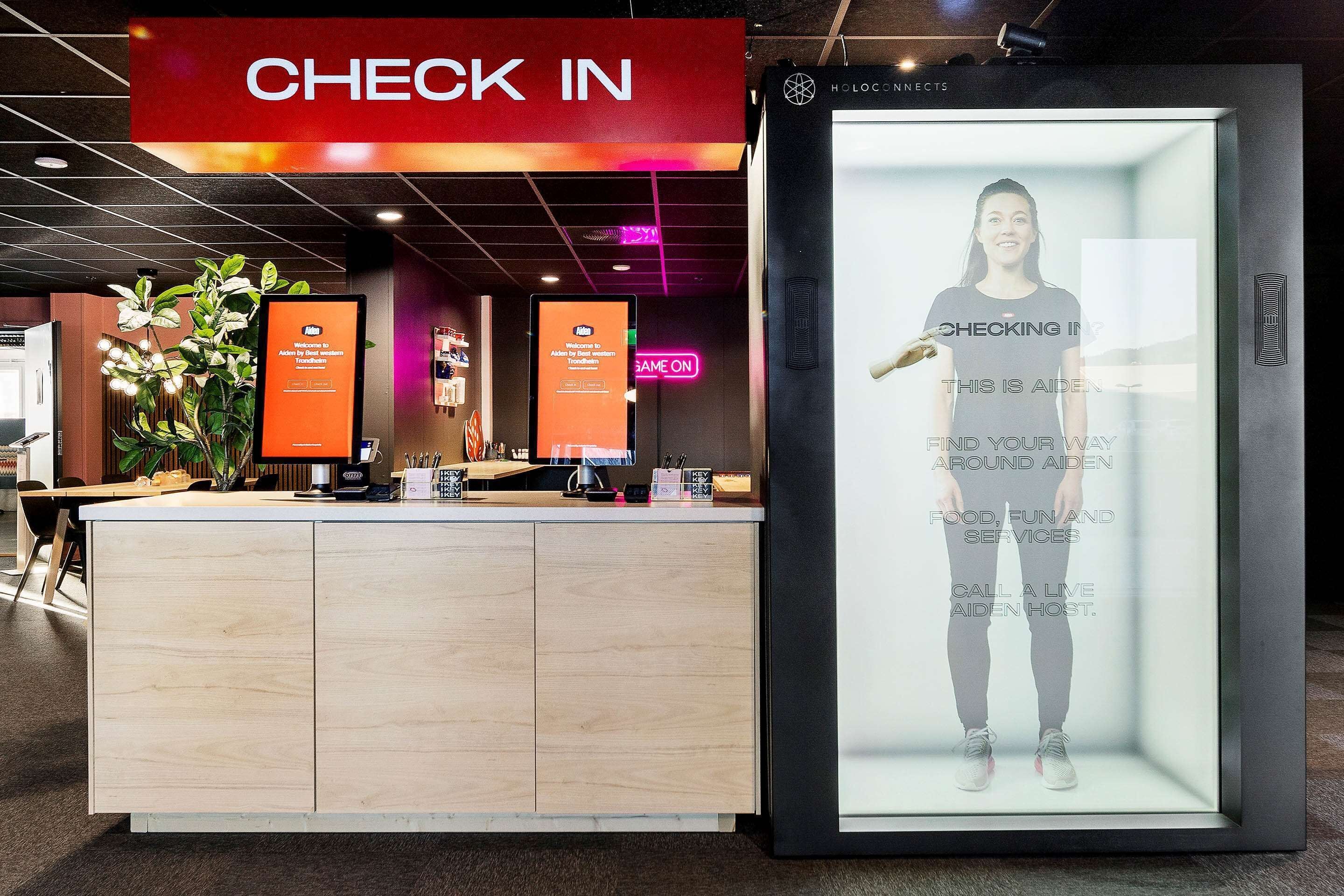 A reception area with a 'CHECK IN' sign, a digital screen displaying a welcome message, and a framed photo of a person with text overlay.