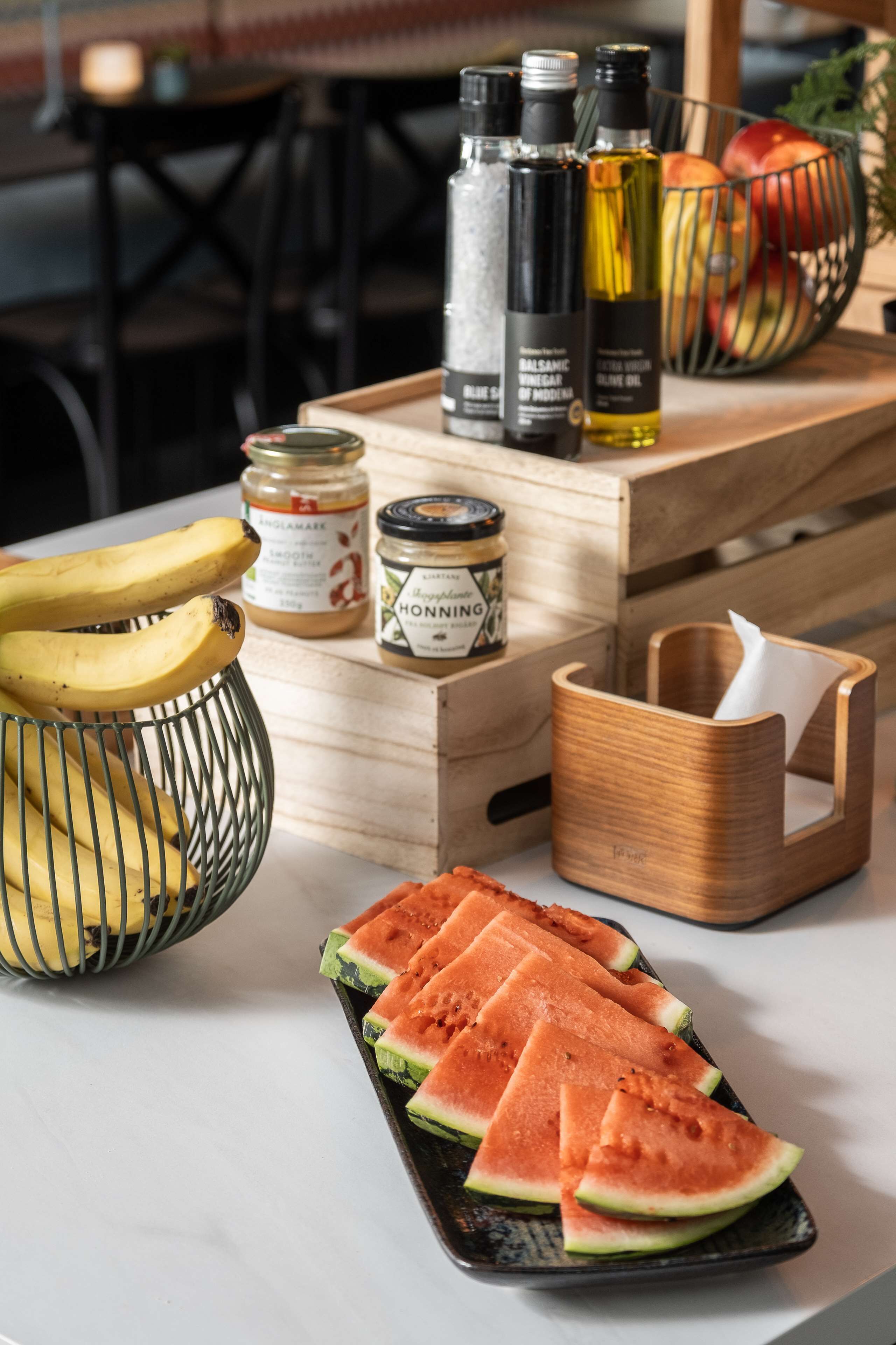 A wooden table with a tray containing sliced watermelon, a basket of bananas, and jars of honey and mustard.