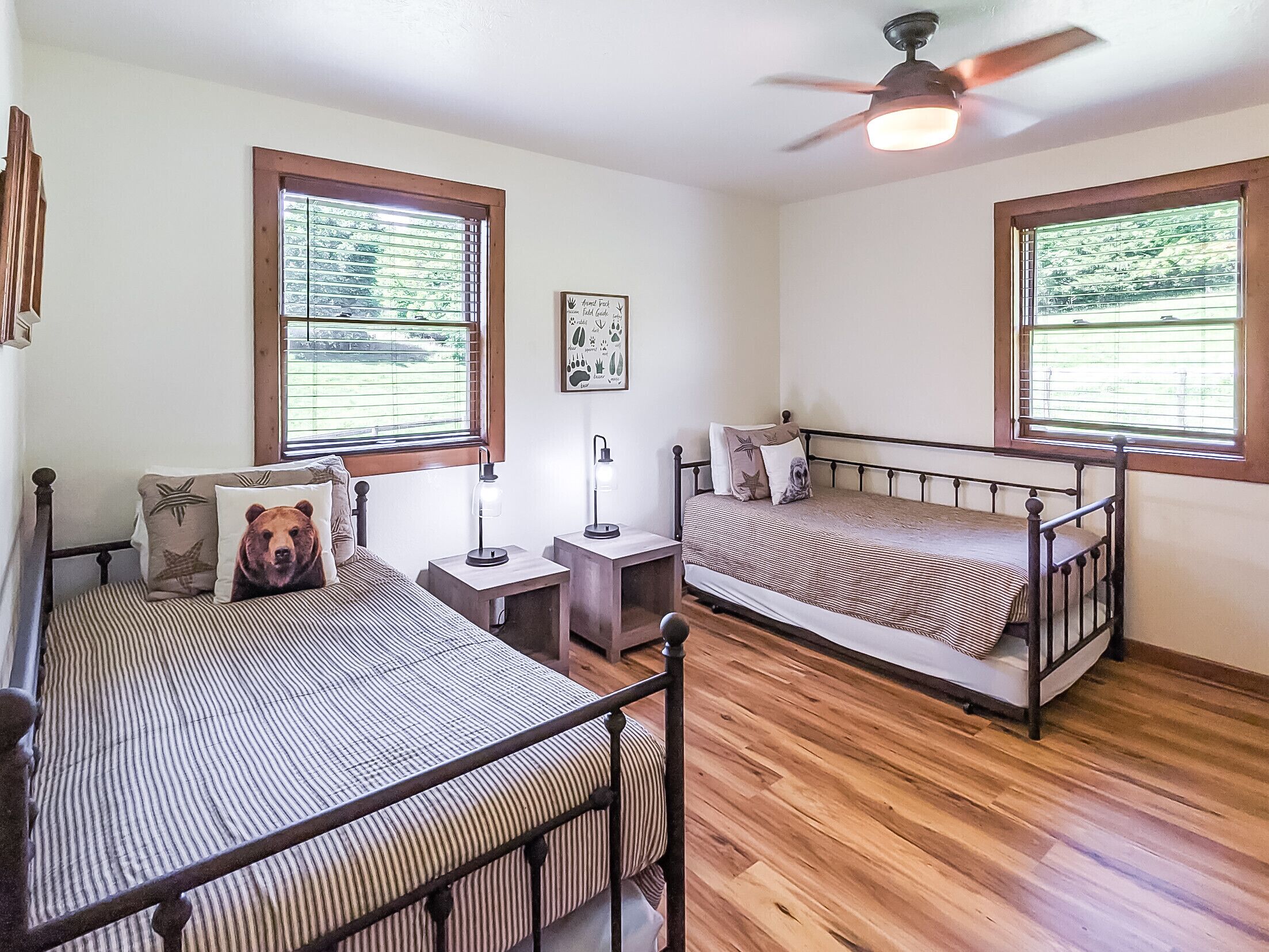 A room with two beds, wooden flooring, and windows with blinds.