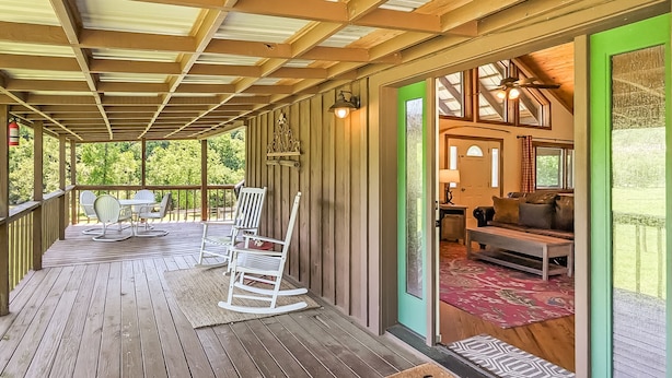 A wooden deck with outdoor seating, a welcome mat, and a view into a cozy interior with a sofa and lamp.