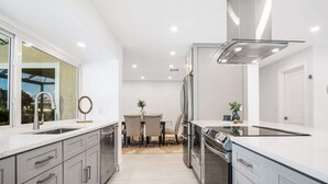 A modern kitchen with stainless steel appliances, a large window, and a dining area with a table and chairs. - Luxury Modern Waterfront 3BR Villa for 8, Heated Pool, Walk to Beach, Top Dining (Marco Island)