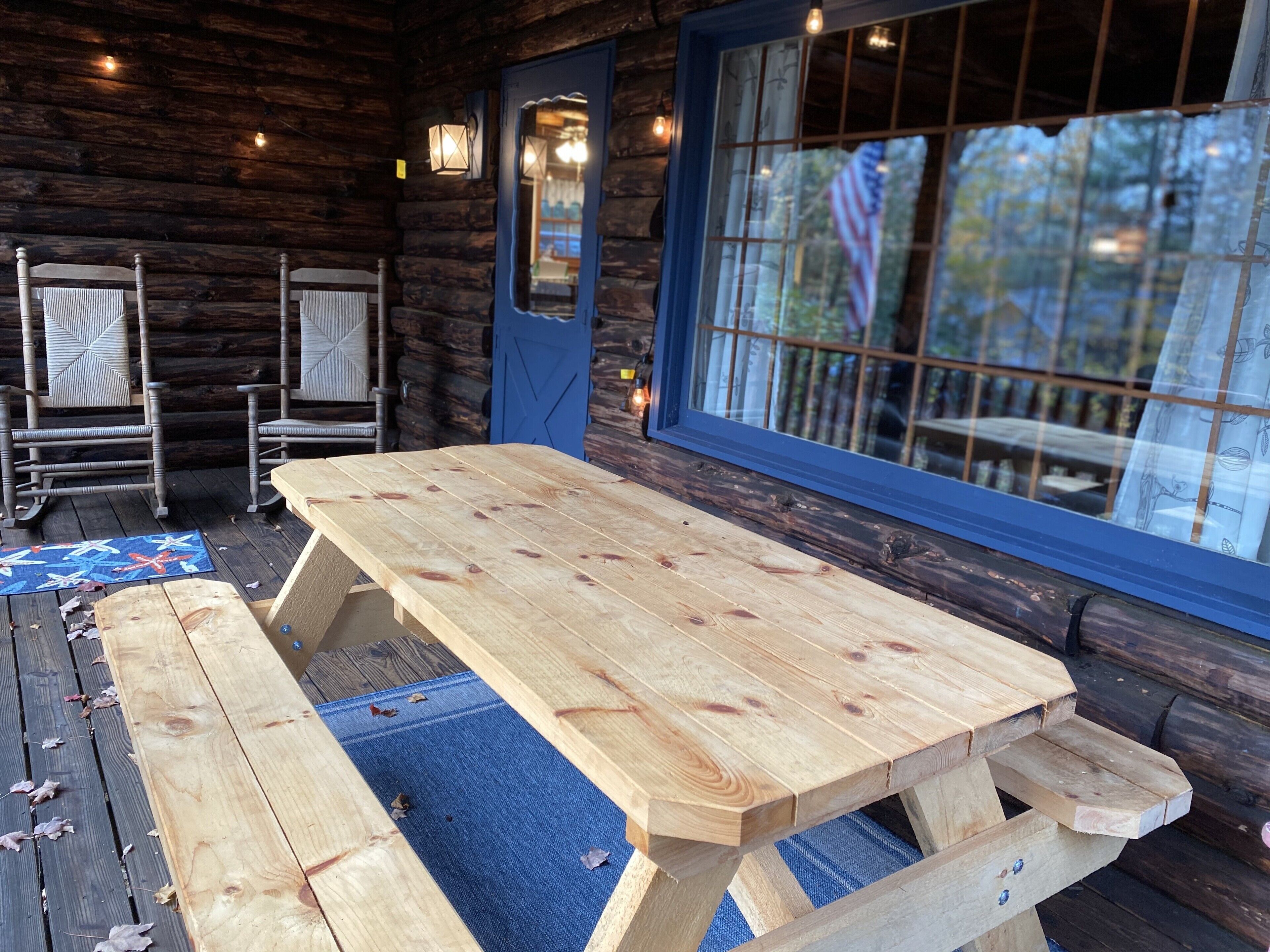 Eine Holzterrasse mit Picknicktisch und Stühlen, eine blaue Tür und ein Fenster mit Blick auf Bäume.