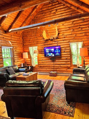 Interior de una cabaña de troncos con chimenea, sofás de cuero, un televisor de pantalla plana y una decoración rústica.