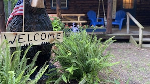 A wooden bear statue holding a welcome sign, surrounded by ferns and flowers, with a rustic wooden building and outdoor seating in the background.