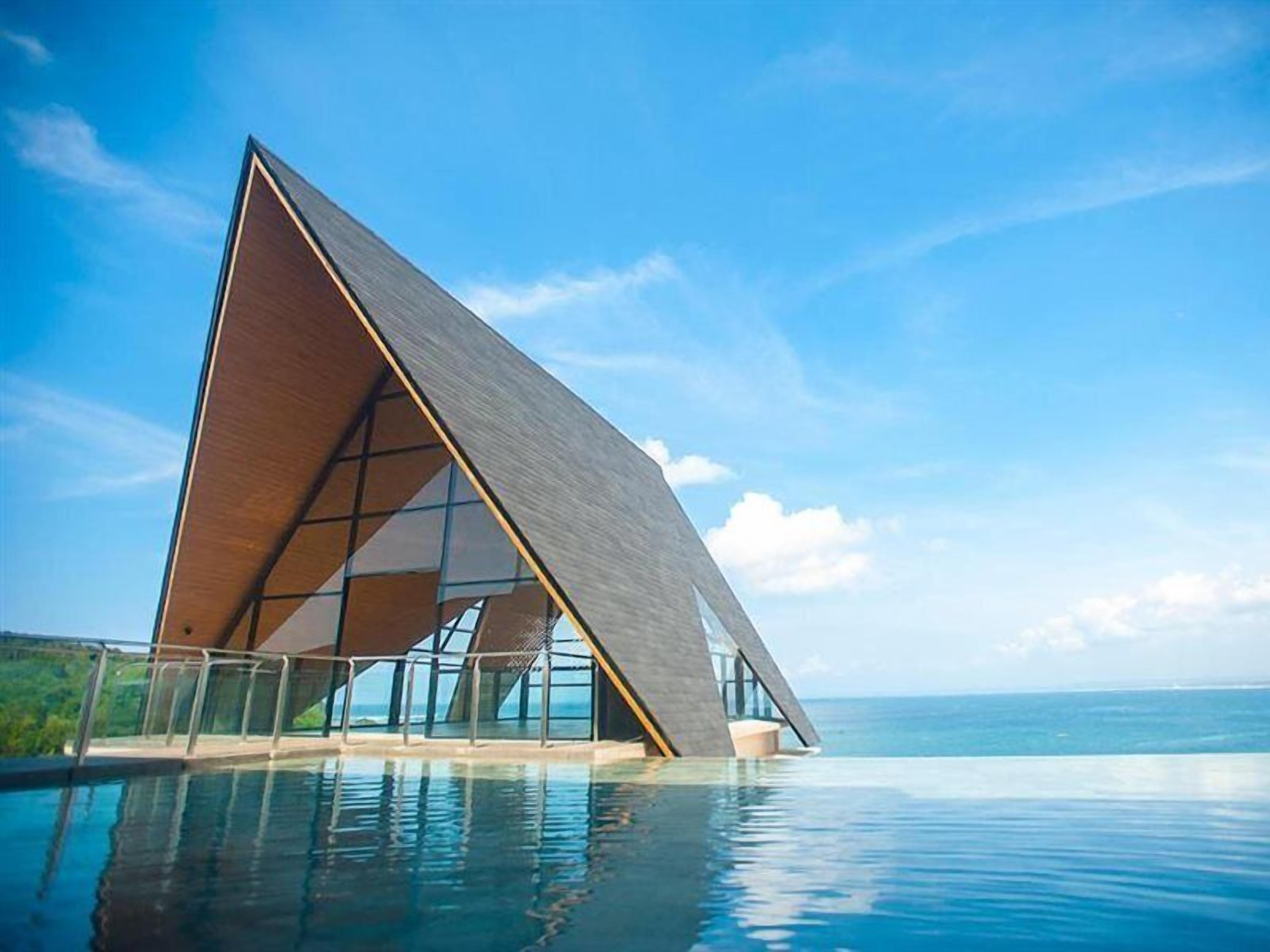 Un edificio moderno con tetto triangolare sopra una piscina a sfioro, con vista su un cielo azzurro e l'oceano.