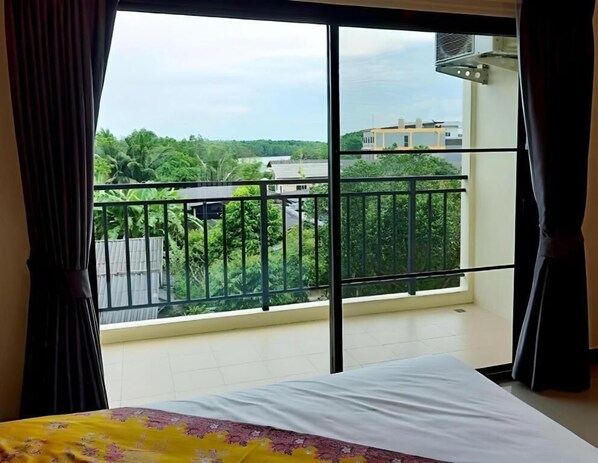 Una habitación con cama, ventana con vista a zonas verdes y edificios, y balcón con barandilla.