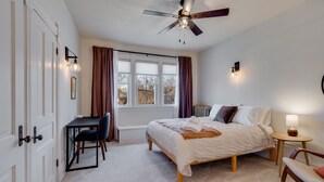 Ein Schlafzimmer mit einem Bett, einem Schreibtisch, einem Stuhl, einem kleinen Tisch mit Lampe und einem Deckenventilator.