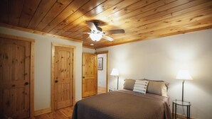 A bedroom with a wooden ceiling, a bed with a dark blanket, two bedside lamps, and wooden doors. - Lakefront Cabin with Hot Tub! (Brookport)