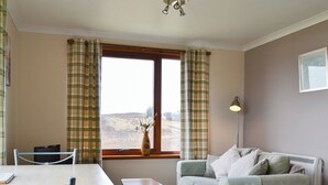 Ein Zimmer mit einer Couch, einem Tisch, einem Sessel, einem Fenster mit Vorhängen, einer Lampe und einer Heizung.