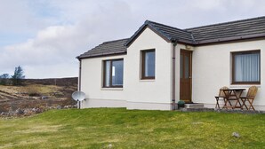 Ein eingeschossiges Haus mit einem geneigten Dach, einer Satellitenschüssel und einer kleinen Terrasse mit Holzstühlen.