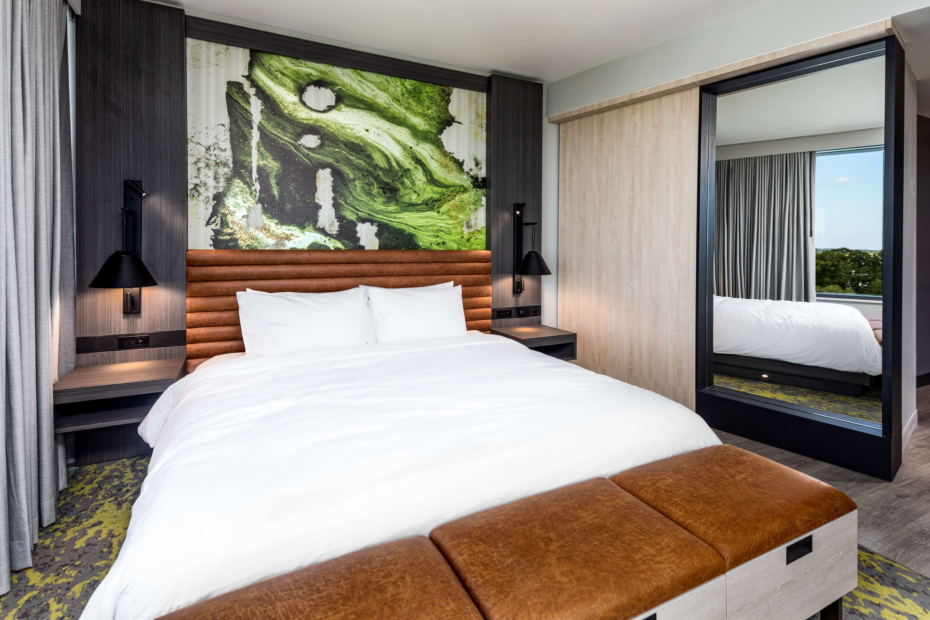 A modern hotel room with a large bed, a wooden headboard, a large mirror, and a view of the outdoors.