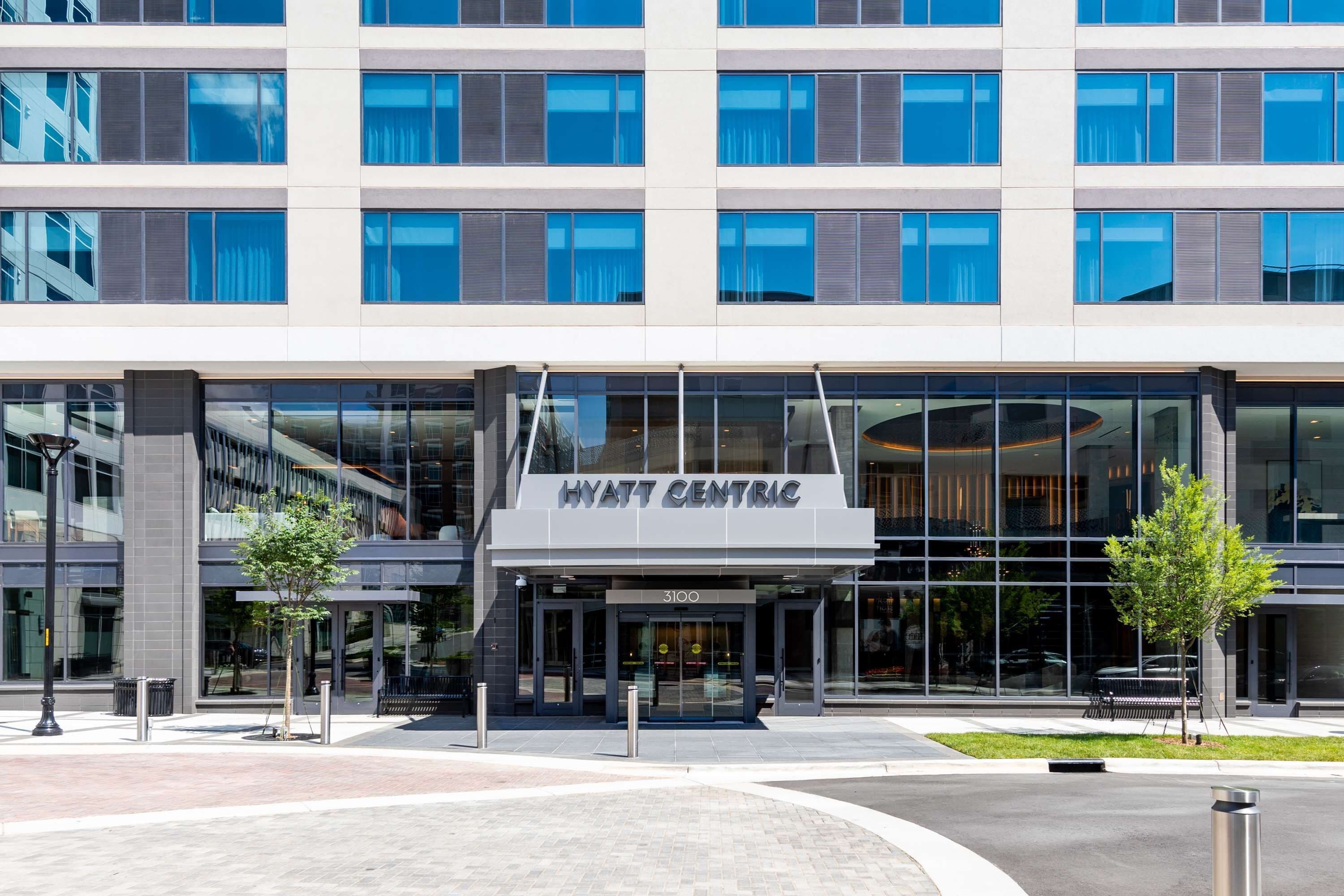 Un hotel moderno con fachada de vidrio, identificado como "HYATT CENTRAL", que cuenta con una entrada destacada y un área ajardinada con árboles.