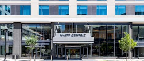 Un hotel moderno con fachada de vidrio, identificado como "HYATT CENTRAL", que cuenta con una entrada destacada y un área ajardinada con árboles.