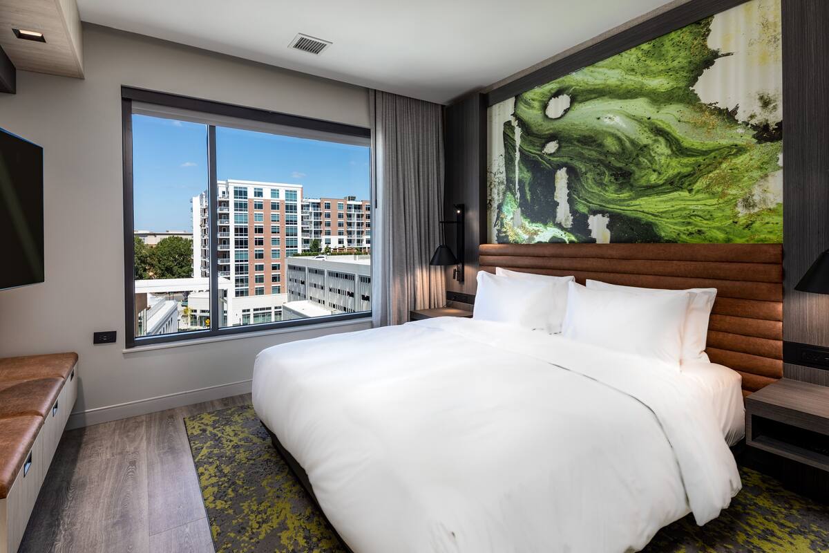 A modern hotel room with a large bed, a view of the cityscape, a wooden headboard, and a large abstract painting.