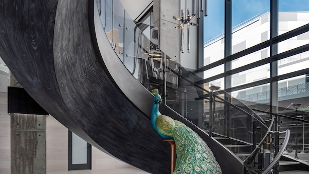 Una escalera de caracol con diseño de pluma de pavo real en la base, luminarias modernas y grandes ventanales con vista al cielo.